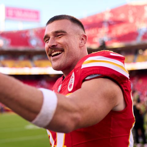 travis kelce smiles as he holds a phone out in front of his face while standing inside a football stadium on the field, he wears a red kansas city chiefs jersey
