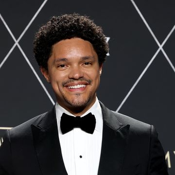 trevor noah smiles at the camera, he wears a black tuxedo and tie with a white collared shirt and stands in front of a black background