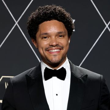 trevor noah smiles at the camera, he wears a black tuxedo and tie with a white collared shirt and stands in front of a black background