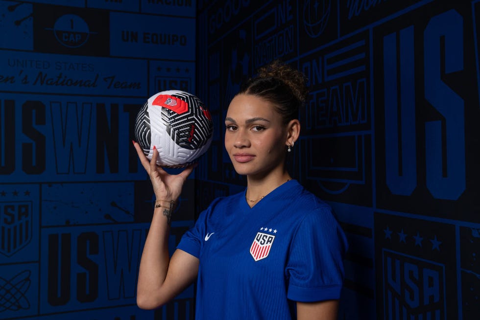 trinity rodman wearing her blue us soccer jersrey and holding a soccer ball in her right hand