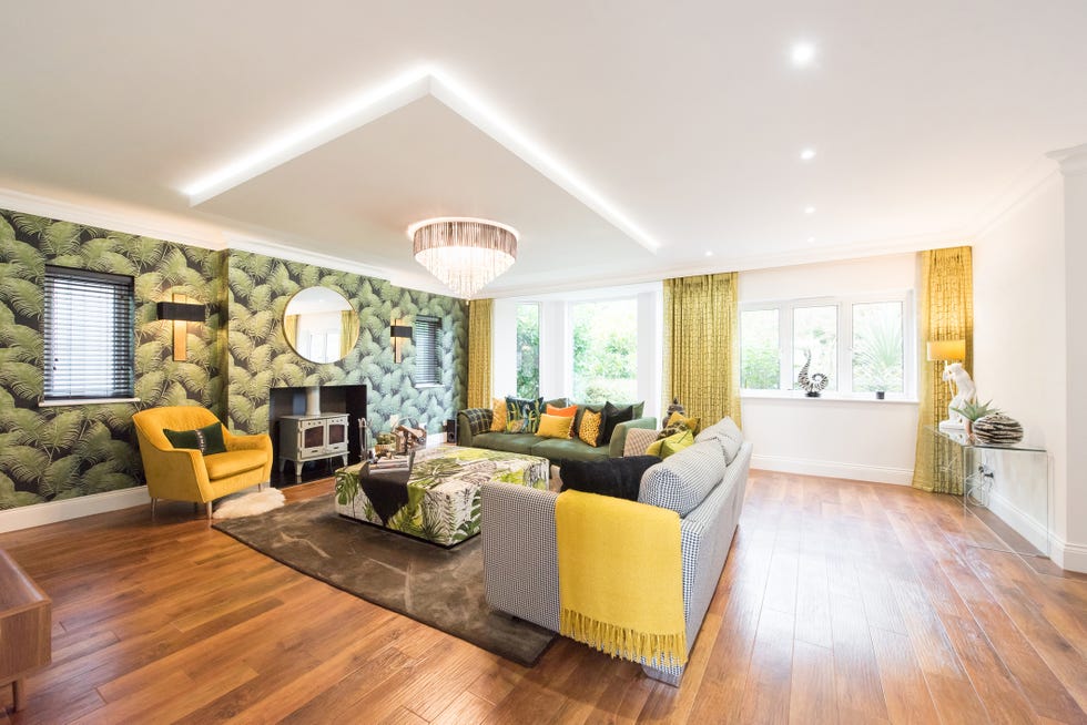 a general interior view of a large lounge with tropical palm leaf print wallpaper, wood burner stove fireplace, two sofas, armchair, chandelier, coffee table, dropped ceiling box with led lighting, rug, hardwood flooring, black and gold wall lights