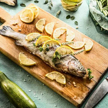 raw trout fish on cutting board stuffed with herbs and lemon slices on light blue kitchen table background with ingredients real cooking , step by step