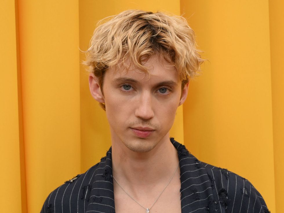 troye sivan standing in front of a yellow curtain wearing a pinstripe suit