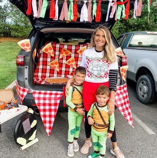 two ninja turtle kids and a pizza delivery mom stand in front of a tmnt themed trunk or treat setup