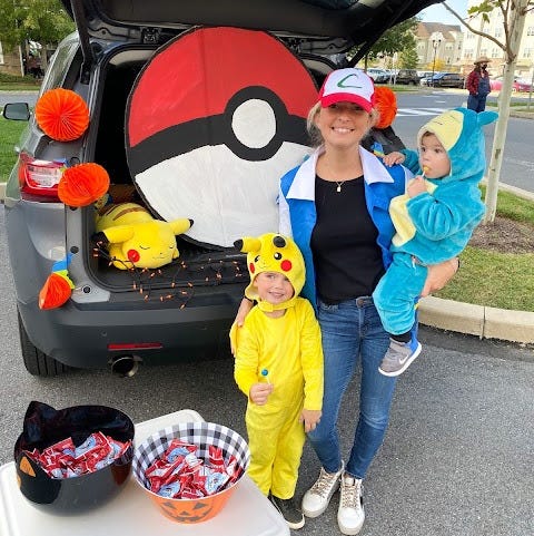 a kid dressed as pikachu, a kid dressed as snorlax, a mom dressed as ash stand in front of a pokemon themed trunk or treat setup