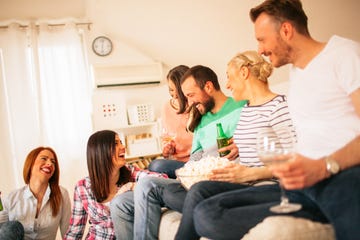 group of laughing friends sitting on couch and floor at home drinking beer and eating popcorn