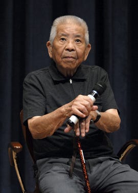 tsutomo yamaguchi sits in a chair and holds a microphone, he looks out with a neutral expression on his face, he wears a black polo shirt and watch