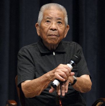 tsutomo yamaguchi sits in a chair and holds a microphone, he looks out with a neutral expression on his face, he wears a black polo shirt and watch