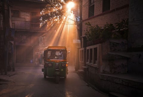 Deze foto is genomen in Jaipur tijdens het gouden uur Je kunt goed zien waarom de inwoners dit de Roze stad van India noemen Het zachte zonlicht aan het einde van de middag dringt door tot elke hoek van de stad waardoor alle kleuren rood en roze extra opvallen Een taxi die door de walm van stofdeeltjes van ander verkeer rijdt geeft dit beeld een betoverende mysterieuze sfeer