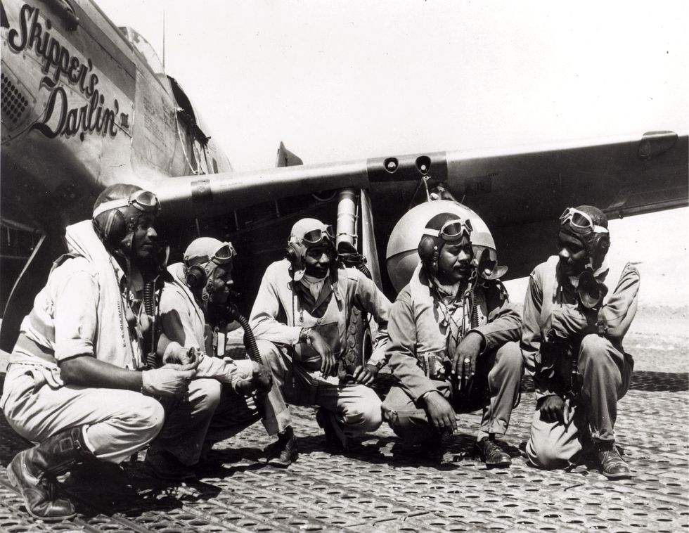 five pilots in uniform talk to each other while kneeling in front of the wing of a military airplane