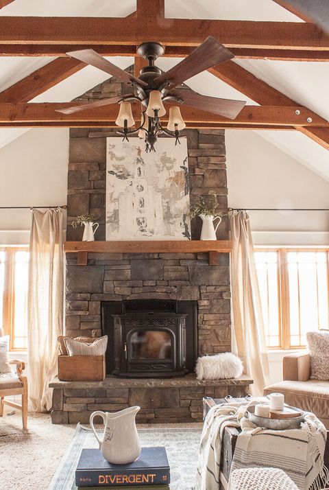 farmhouse style living room with stone fireplace white pitchers used as decor on the mantle, an old trunk is used as a coffee table with a thrown and candles in an old tin tray on it the ceiling has exposed wooden beams and the walls are white