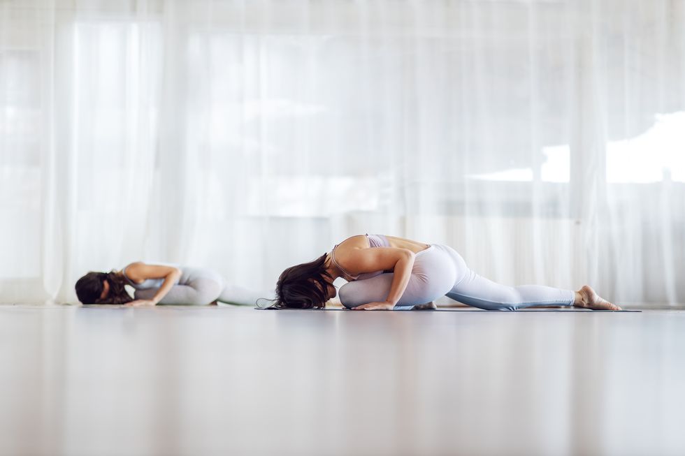 Two attractive fit yogi girls in Sleeping Swan yoga pose. Yoga studio interior.