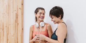 two friends women at the gym talking happily, listening to music and using smart phone. Technology, sports and friendship concept