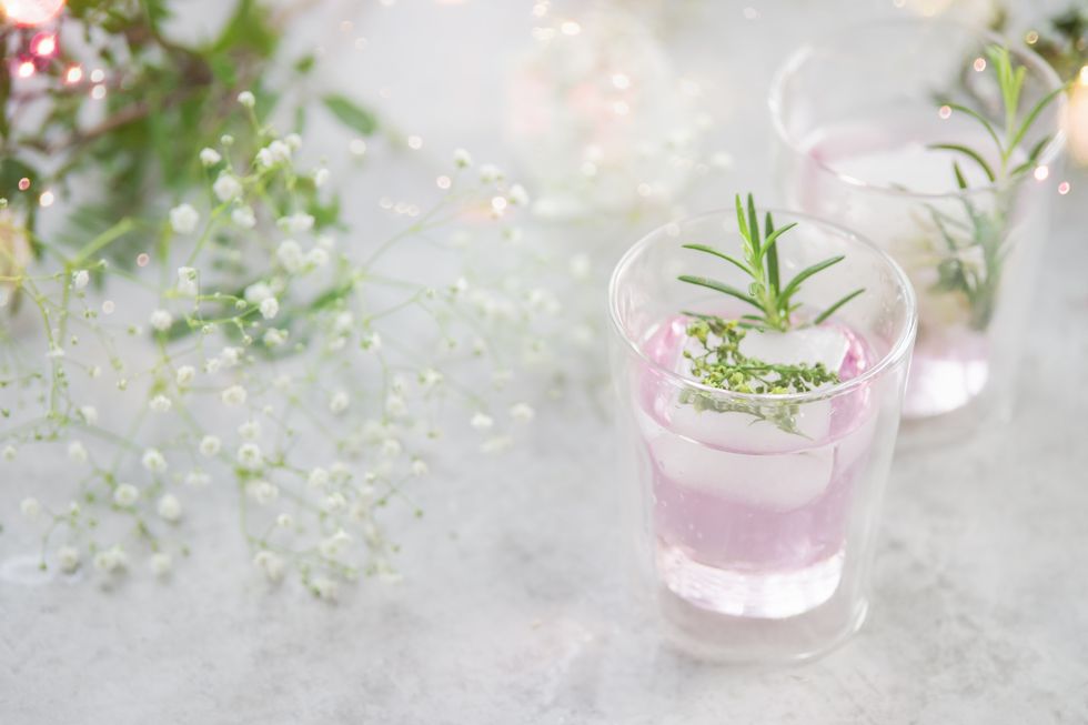 two glasses with pink drink and ice cubes with herbs and green leaves at grey concrete kitchen table with small white flowers and bokeh light effects