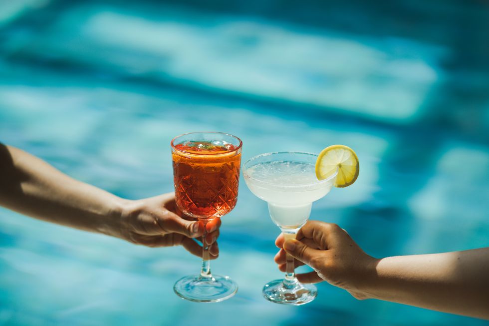 two hands toasting with glasses spritz and margarita cocktails on background of swimming pool cheers