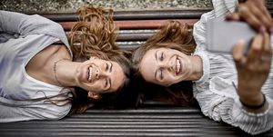 Two happy young women lying on a bench using cell phone