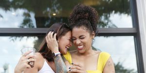 two millennial women, one caucasian with shoulder length hair and the other mixed race with curly hair, laugh while they hug each other outdoor near a building, wearing casual summer clothing