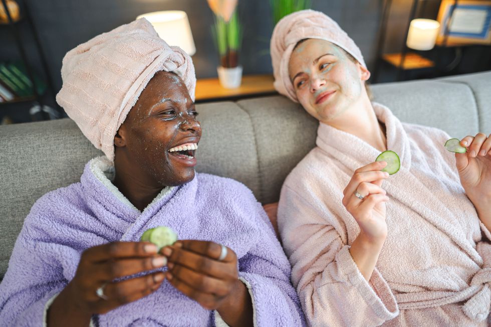 two multiracial female friends with face masks removing cucumber circles from eyes