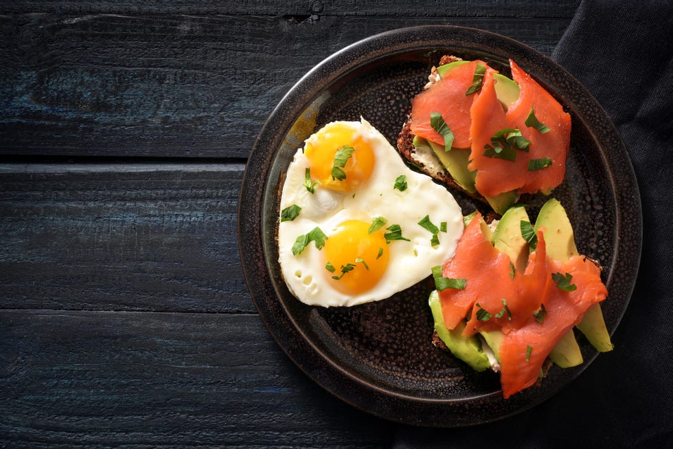 two sandwiches with avocado and smoked salmon and two fried eggs in one heart shape, romantic valentines day meal for a couple on a dark blue wooden table, copy space, high angel view from above