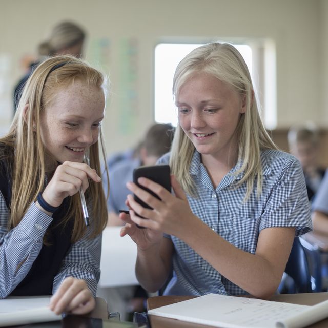 Two smiling schoolgirls in classroom looking at cell phone