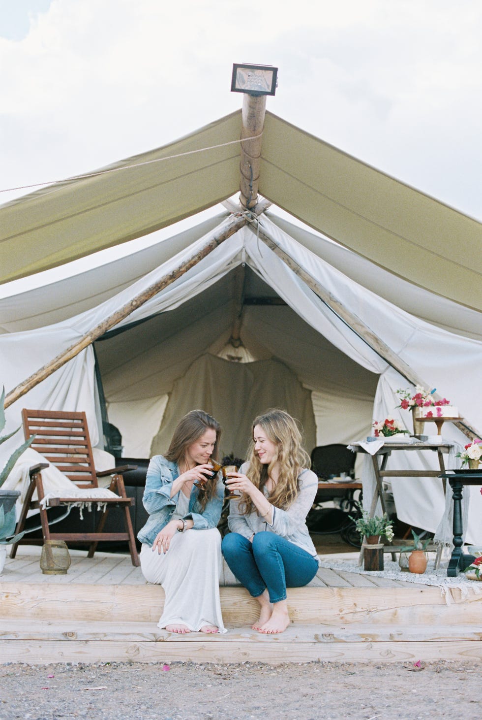 two smiling women sitting outside a large tent laughing and having a glass of wine