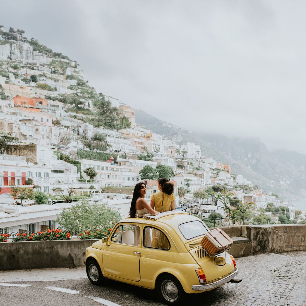 two woman stand in a vintage yellow fiat, and admire the view of positano, italy