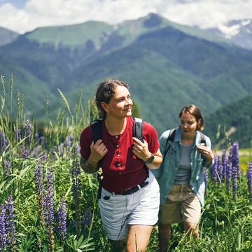 two women hiking through a field of purple lupine flowers. mountains in the background on a sunny day.