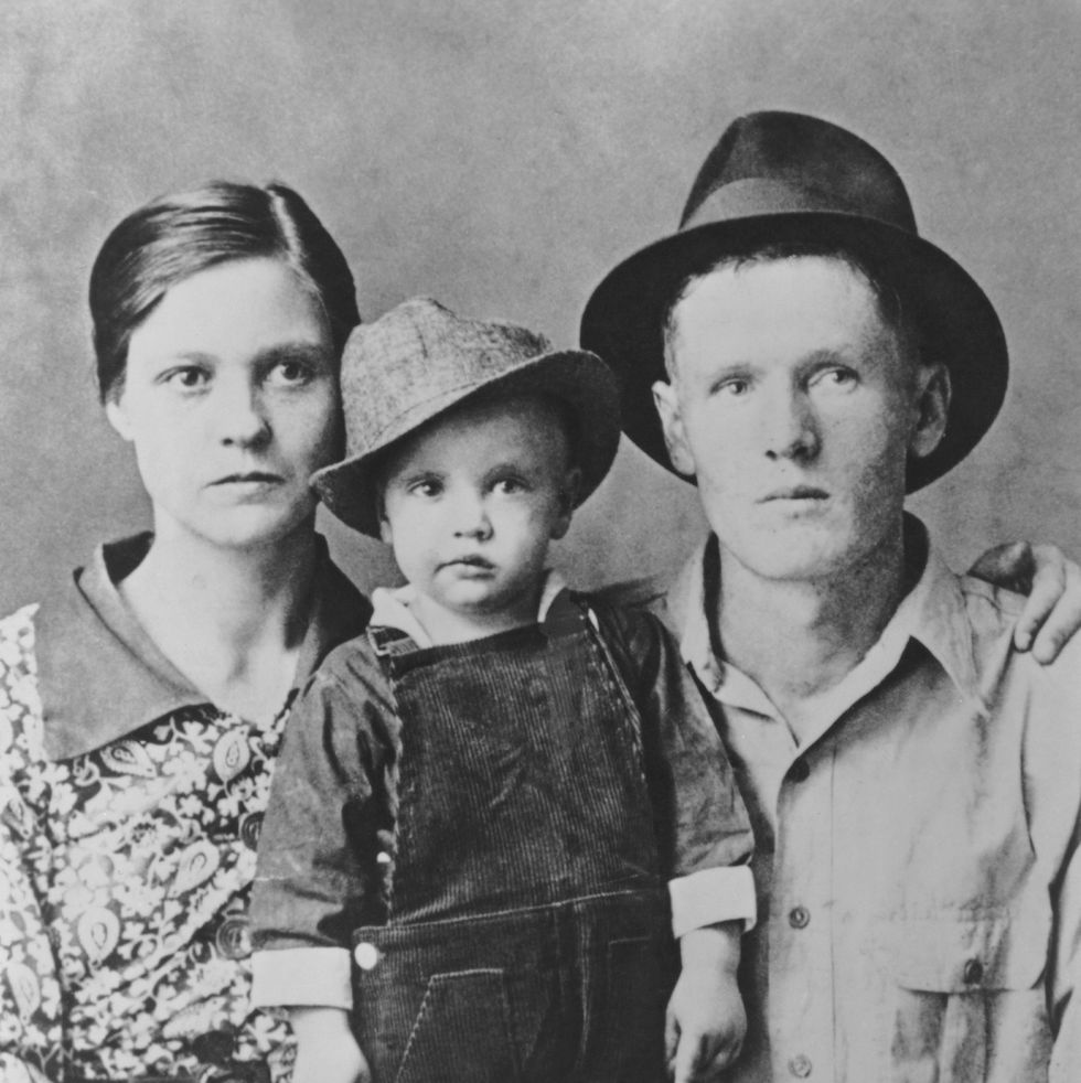 a black and white photo of a man and woman posing with a child, who wears overalls and a fedora
