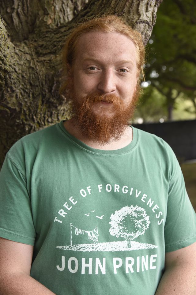 tyler childers smiles at the camera while standing in front of a tree, he wears a green graphic tshirt