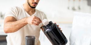 unrecognizable young sporty guy making protein shake at table in kitchen, closeup cropped view of millennial bodybuilder using sports nutrition supplement, preparing cocktail indoors