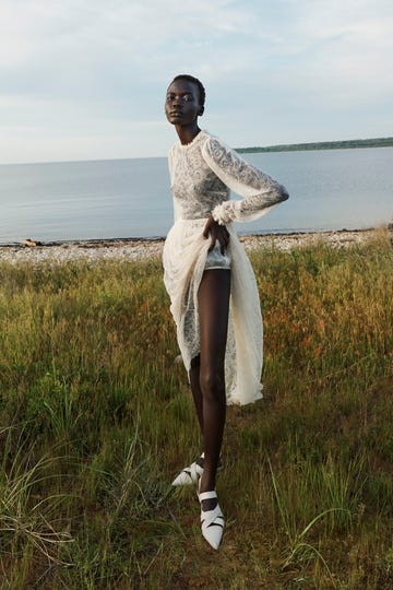 a person in a white dress standing in a grassy field by a body of water