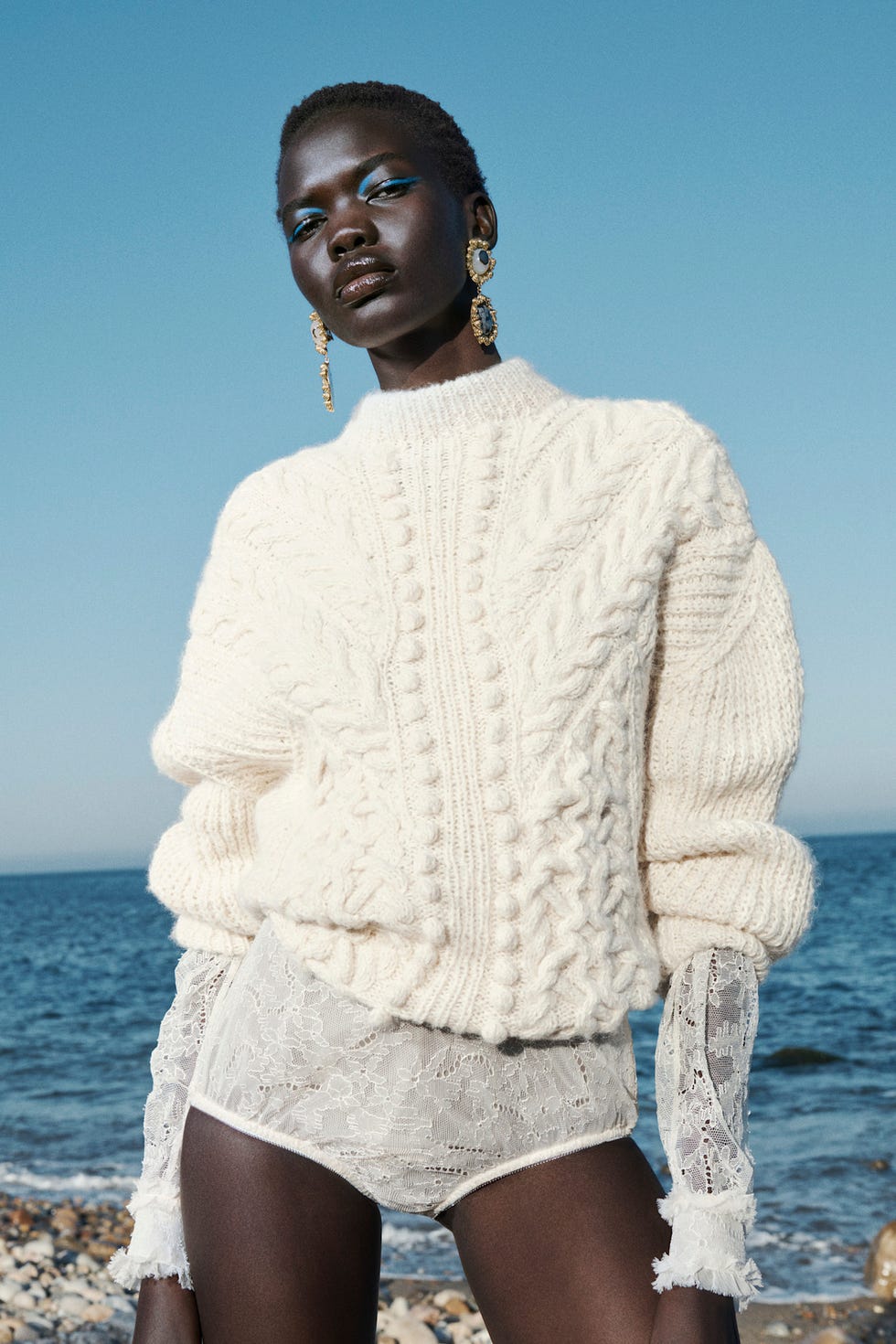 Model wearing a thick white cableknit sweater and lace shorts near the ocean
