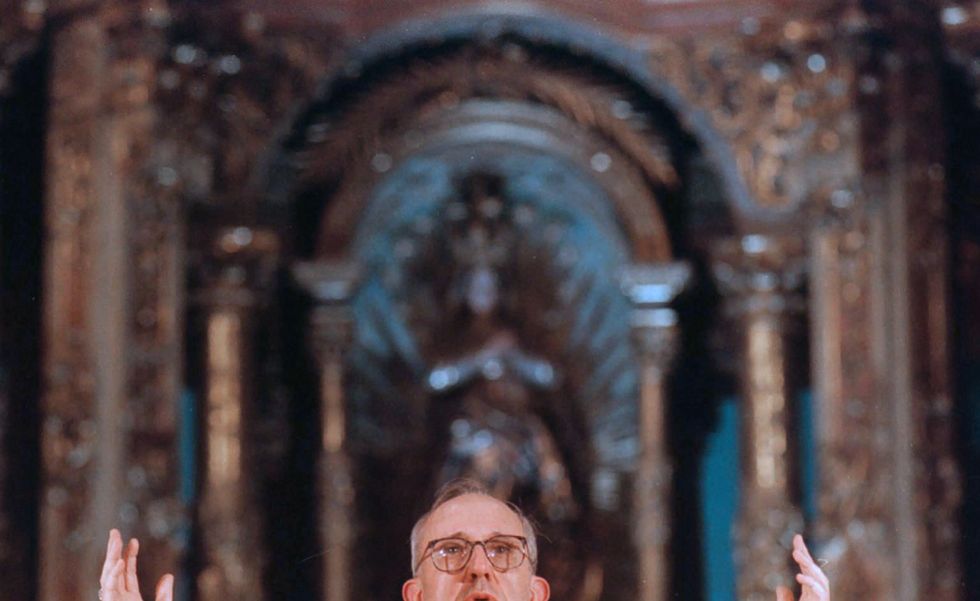 jorge bergoglio aka pope francis stands at a pulpit and raises both hands up, he wears white priest robes with a red center panel