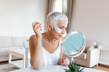 under eye masks for puffiness, wrinkles, dark circles eye patches concept smiling senior gray haired woman in white shirt, applying silver eye patches while looking in the mirror