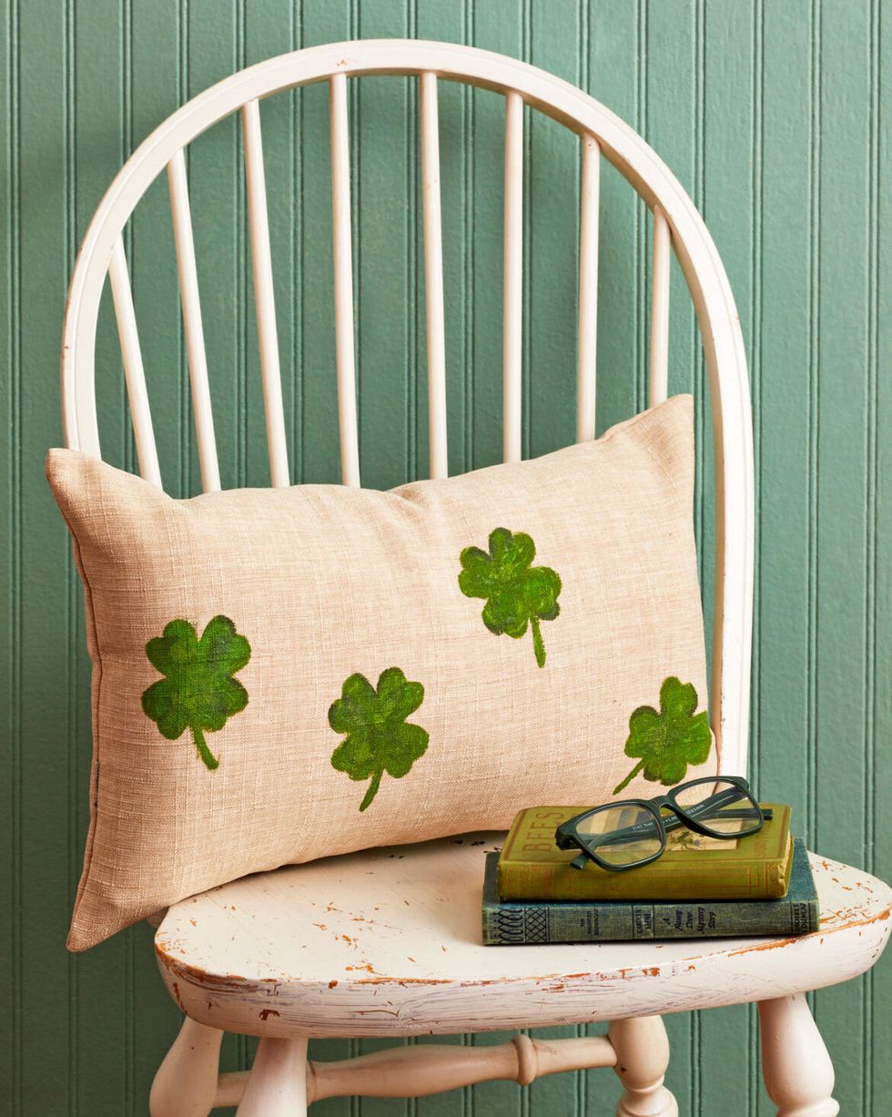a linen pillow with green four leaf clovers painted on the front resting on a white chair behind a small stack of green books