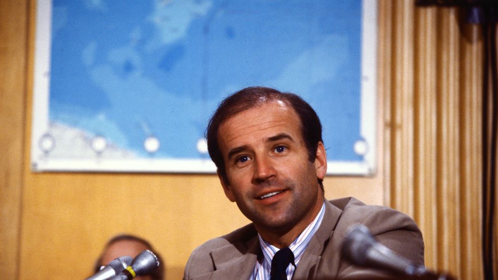 joe biden smiles as he sits at a table with a microphone in front of him, he wears a suit jacket tie and striped collared shirt