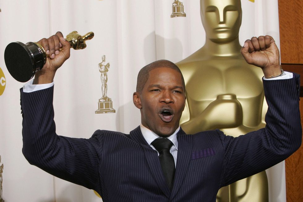 jamie foxx wearing a black suit and tie, pumping both his fists into the air, holding an oscar statuette in his right hand, and standing in front of a large oscar statue