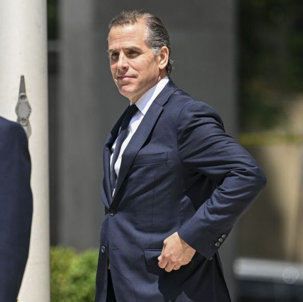 hunter biden stands outside a building wearing a navy blue suit jacket and lifts his arm to reach his pocket