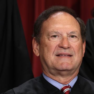 samuel alito smiles slightly at the camera, he wears black robes over a light blue collared shirt and red tie with a white and blue stripe