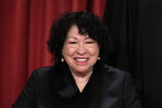 sonia sotomayor wears her black supreme court justice robes, dangling pearl earrings, and red lipstick, she smiles at the camera, and her curly hair frames her face down to her neck