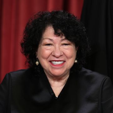 sonia sotomayor wears her black supreme court justice robes, dangling pearl earrings, and red lipstick, she smiles at the camera, and her curly hair frames her face down to her neck