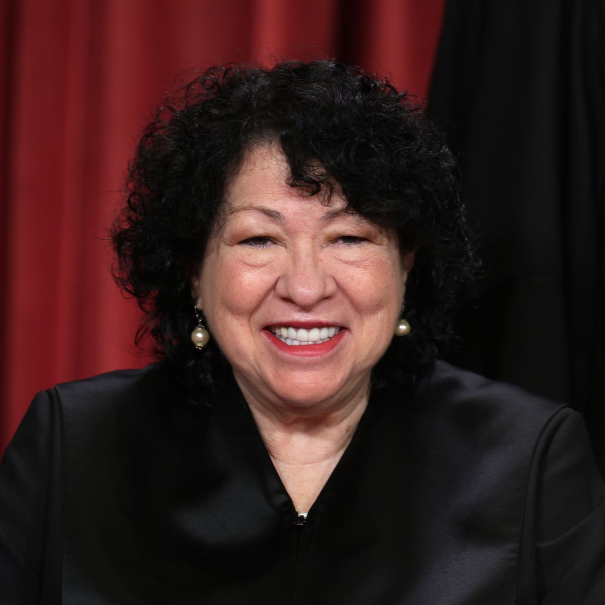 sonia sotomayor wears her black supreme court justice robes, dangling pearl earrings, and red lipstick, she smiles at the camera, and her curly hair frames her face down to her neck
