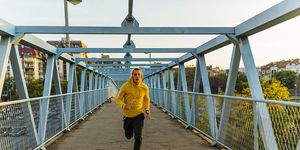 young strong athlete in yellow hoodie jogging outdoors at the bridge and listening to music with headphones, male runner working out on the city bridge sportsman do physical exercises