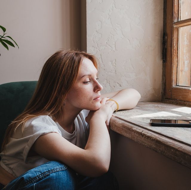 upset redhead teen girl sitting by window looking at phone waiting call or message