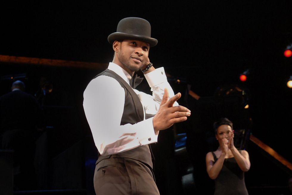 usher wearing a black vest, white shirt, black pants, and black hat, performing on stage with another actor behind clapping behind him
