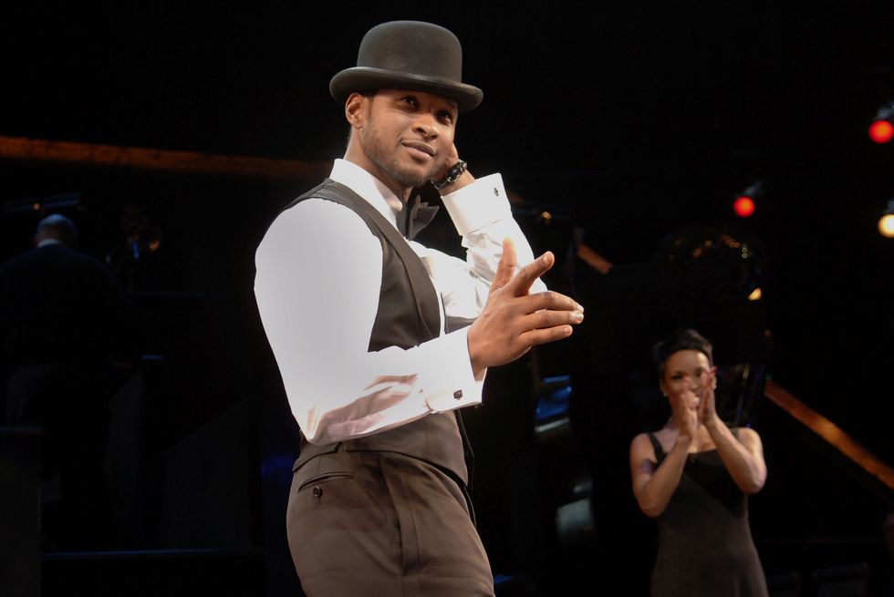usher wearing a black vest, white shirt, black pants, and black hat, performing on stage with another actor behind clapping behind him