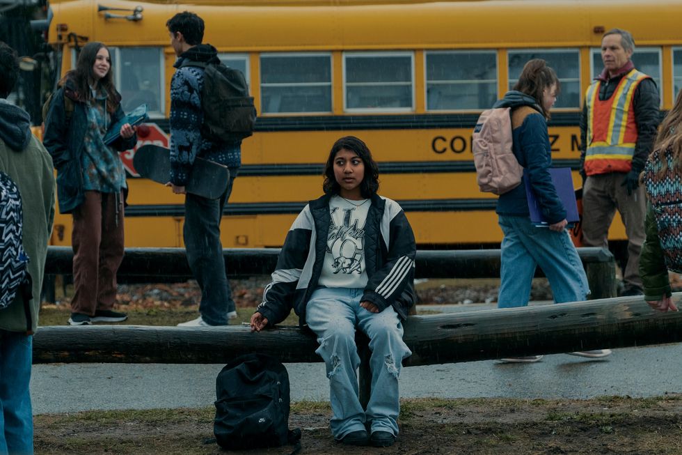 a teenage girl sits on a log railing and looks to the left, a backpack sits on the ground to the left, fellow students and a yellow school bus are in the background