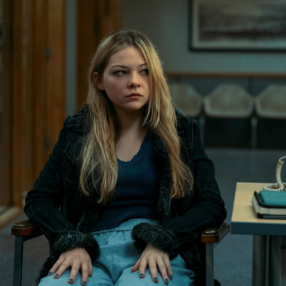 a girl sits in an armchair and looks to the right, she wears a sweater, a scoop neck shirt and pants, a desk with a phone and notebooks is on the right