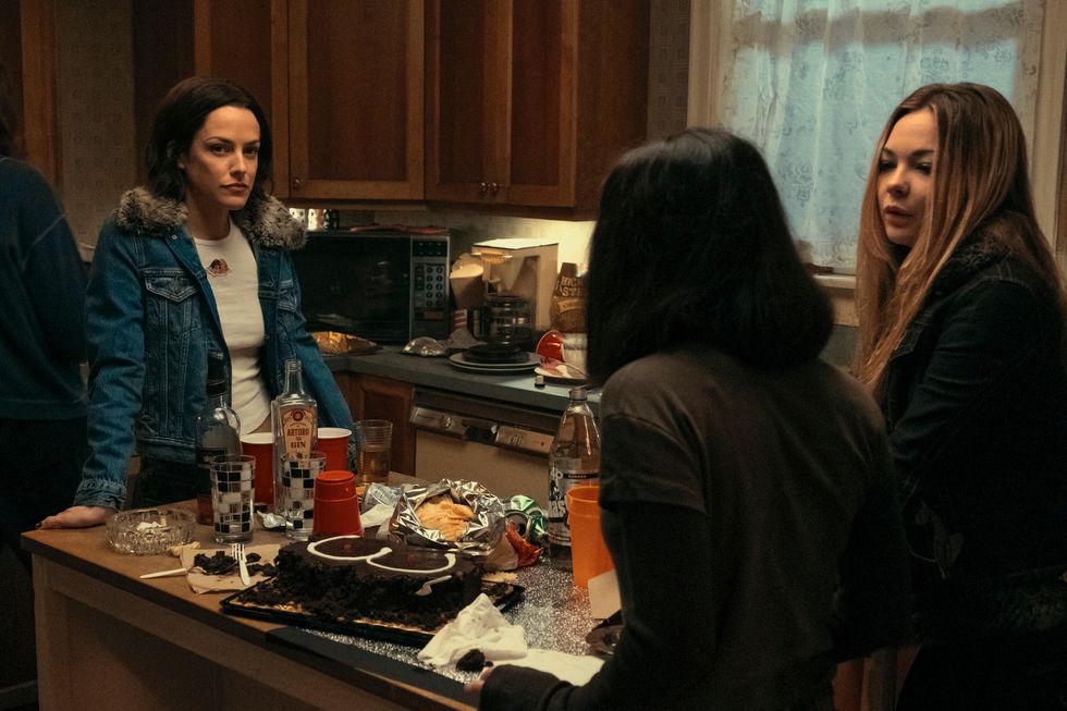 riley keough leans against an island in a kitchen that has a cake, cups, drinks, and other items on it, on the other end of the island are two teenage girls facing her