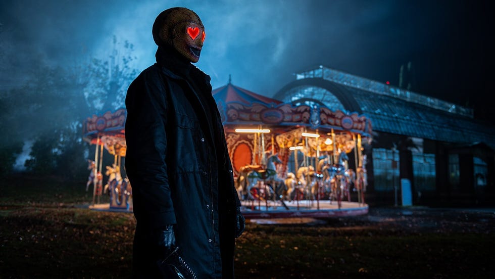 a dark figure wearing a mask with glowing heart eyes stands near a carousel at night
